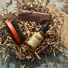 Load image into Gallery viewer, A bottle of Raw By Nature Beard Oil, a brush, and a comb, all displayed on a bed of brown cardboard packing material.