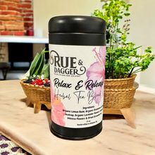 Load image into Gallery viewer, Rue & Dagger herbal tea blend container on a wooden surface with plants in the background