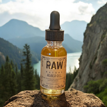 Load image into Gallery viewer, A bottle of Raw By Nature Beard Oil with a dropper, placed on a rocky surface with a scenic mountain backdrop.