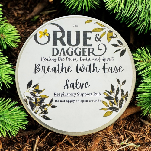 Round container of 'Rue & Dagger' salve with greenery in the background