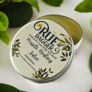 Open Tin of Rue & Dagger Muscle Healing Salve on white surface with leaves