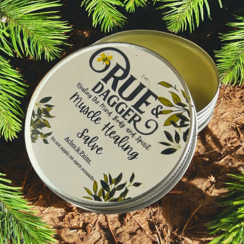 Open tin of Rue & Dagger Muscle Healing Salve on a natural background with greenery.