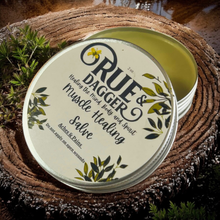Load image into Gallery viewer, Tin of Rue &amp; Dagger Muscle Healing Salve on a wooden surface with plants around

