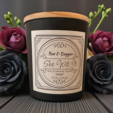 Load image into Gallery viewer, Candle labeled 'Rue &amp; Dagger She Wit It' with decorative roses in the background
